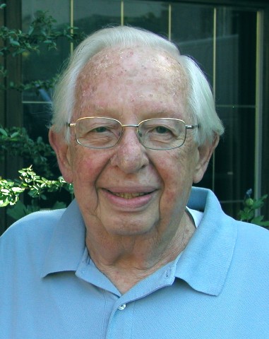 Frank Alper in a garden, wearing a soft blue shirt, smiling gently toward the camera.